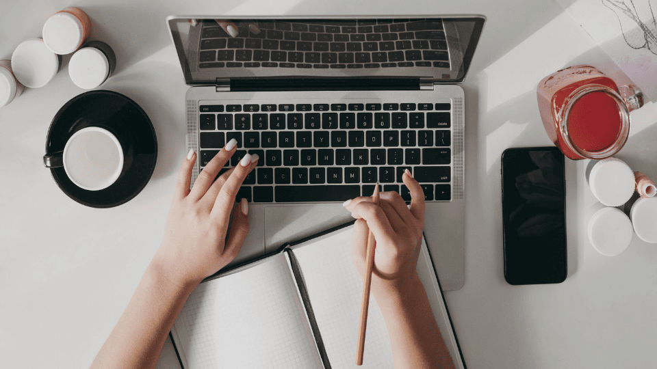 Person content writing on a laptop, with a notebook, coffee, and phone on the desk.